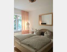 Helles Schlafzimmer mit Doppelbett in Holzoptik, grauer Bettwäsche und mehreren Kissen, daneben ein Nachttisch mit Lampe und ein großer Spiegel an der Wand. Links ein Fenster mit halbtransparenten rosafarbenen Vorhängen und Blick ins Grüne.