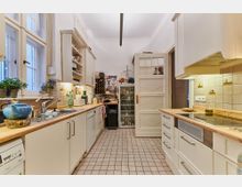 Helle Küche mit cremeweißen Schränken, Holzarbeitsplatten und Fliesenboden. An den Wänden befinden sich beige Wandfliesen mit kleinen Mustern, es gibt sichtbare Küchengeräte, eine Spüle und Regale sowie eine offene Tür im Hintergrund.