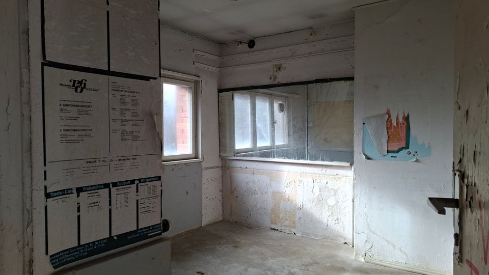 Kleiner, leerer Innenraum mit abgenutzten weißen Wänden, Betonboden und zwei Fenstern; eine große Glasscheibe trennt einen Bereich wie einen alten Kassen- oder Empfangsschalter ab. An der linken Wand hängen mehrere Plakate und Aushänge, rechts ist ein teilweise abgerissenes Wandbild zu sehen.