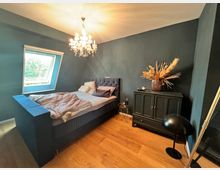 Ein Schlafzimmer mit dunkelblauen Wänden, einem großen gepolsterten Bett und Holzboden. Ein Kronleuchter hängt von der Decke, ein modernes Schrägfenster lässt Tageslicht herein, und eine Kommode mit Trockenblumen steht an der Wand.