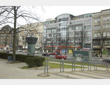 Straßenszene in einer Stadt mit mehrstöckigen Gebäuden, darunter moderne und ältere Fassaden. Im Vordergrund Gehwege, grüne Büsche, eine alte Lüftungs- oder Turmstruktur und Straßenverkehr mit parkenden und fahrenden Autos.