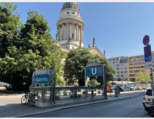 U-Bahnhof-Eingang „Stadtmitte“ mit dekorativem Metallgeländer im Vordergrund, umgeben von Bäumen und urbaner Umgebung; im Hintergrund die Kuppel des Französischen Doms auf dem Berliner Gendarmenmarkt und moderne Gebäude.