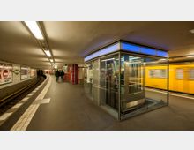 U-Bahnhof mit gekrümmtem Bahnsteig, einem gläsernen Aufzug in der Mitte und einer gelben U-Bahn, die rechts am Bahnsteig einfährt. Die Wände sind mit Werbung versehen, und Deckenleuchten sorgen für eine gleichmäßige Beleuchtung.