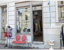 Außenansicht eines kleinen Bekleidungsgeschäfts mit einer Schaufensterscheibe, die Kleidungsstücke und Dekorationen zeigt. Vor dem Laden stehen eine rote Metallbank, ein dekoratives Tischchen mit Keramik sowie eine Schaufensterpuppe mit bunter Kleidung.