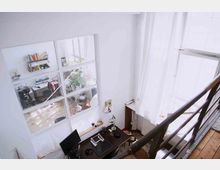 Mehrstöckiges Büro mit Blick von einer Treppe auf einen kleinen Arbeitsbereich mit Schreibtisch, Lampe und Bildschirm. Durch eine Glaswand sind ein angrenzender Raum mit Schreibtischen, einem Regal und Pflanzen zu sehen, während Tageslicht durch große Fenster mit weißen Vorhängen einfällt.