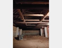 Alter Innenraum eines ehemaligen landwirtschaftlichen Gebäudes mit Backsteinwänden, sichtbaren Holzbalken an der Decke und einem Betonboden. Der Raum wirkt leer, mit verstreuten Stroh- und Schmutzresten sowie einer Palette mit dunklem Material in der Ecke.