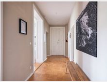 Langer, schlichter Flur in einer Wohnung mit hellbeigen Wänden und Holzboden. Auf der rechten Seite hängt ein großes abstraktes Schwarz-Weiß-Bild, darunter steht eine schlichte Holzbank; links ist ein kleiner gerahmter Druck an der Wand, und am Ende des Flurs befinden sich weiße Doppelflügeltüren.