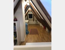 Ein schmaler Dachbodenraum mit sichtbaren Holzbalken und einer Holzfußboden. In der Mitte liegt ein Teppich, links ein Regal mit Spielzeug und rechts einige Aufbewahrungsboxen. Am Ende des Raums befindet sich ein kleines Fenster.