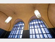 Innenansicht eines Gebäudes mit hohen, bogenförmigen Fenstern aus Gitterglas, einer Decke mit kleinem, geometrischem Muster in Rottönen und hängenden, zylinderförmigen Leuchten. Die unteren Wände sind mit dunkelgrünen Verkleidungen versehen.