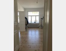 Ein heller, leerer Raum mit hellem Holzfußboden, einem großen Fenster mit weißem Rahmen und Heizkörper darunter. Links steht ein offener, weißer Kleiderschrank mit hängender Kleidung, rechts ist ein Stativ sichtbar.