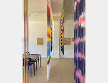 Kunstgalerie mit einer Installation aus farbigen, herabhängenden Fäden, die den Raum in verschiedene Bereiche unterteilen. Im Hintergrund sind weiße Wände mit abstrakten Gemälden und ein Tisch mit schwarzen Stühlen sichtbar.