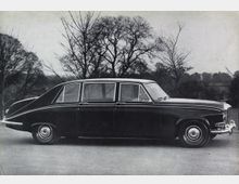 Schwarz-weiß-Fotografie eines klassischen, viertürigen Luxusautos aus den 1960er Jahren, das seitlich auf einer Straße geparkt ist. Im Hintergrund sind kahle Bäume und eine ländliche Landschaft erkennbar.