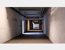 Ein einfacher, schmaler Flur mit weißen Wänden, spärlicher Beleuchtung und freiliegenden Rohren an der Decke. Im Hintergrund ist ein Schild mit der Aufschrift „MUSEUM“ zu sehen.