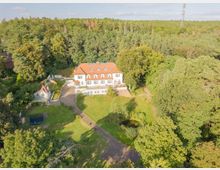 Luftaufnahme einer großen weißen Villa mit rotem Ziegeldach, umgeben von einem weitläufigen Garten mit einem Teich und einem Trampolin. Im Hintergrund ist ein dichter Wald zu sehen, der die ruhige ländliche Umgebung betont.