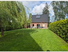 Rasenfläche in einem gepflegten Garten hinter einem Einfamilienhaus aus Klinker mit steilem, dunklem Satteldach und Dachfenstern. Der Garten ist von hohen Hecken und Bäumen eingerahmt und hat eine kleine Terrasse sowie ein schlichtes Schaukelgestell.