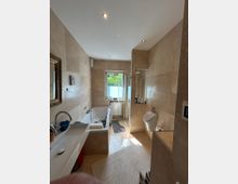 Helles Badezimmer mit cremefarbenen Wand- und Bodenfliesen. Der Raum enthält eine Badewanne, ein Waschbecken, eine Duschkabine und ein wandmontiertes WC; ein Fenster am Ende des Raumes lässt Tageslicht herein.