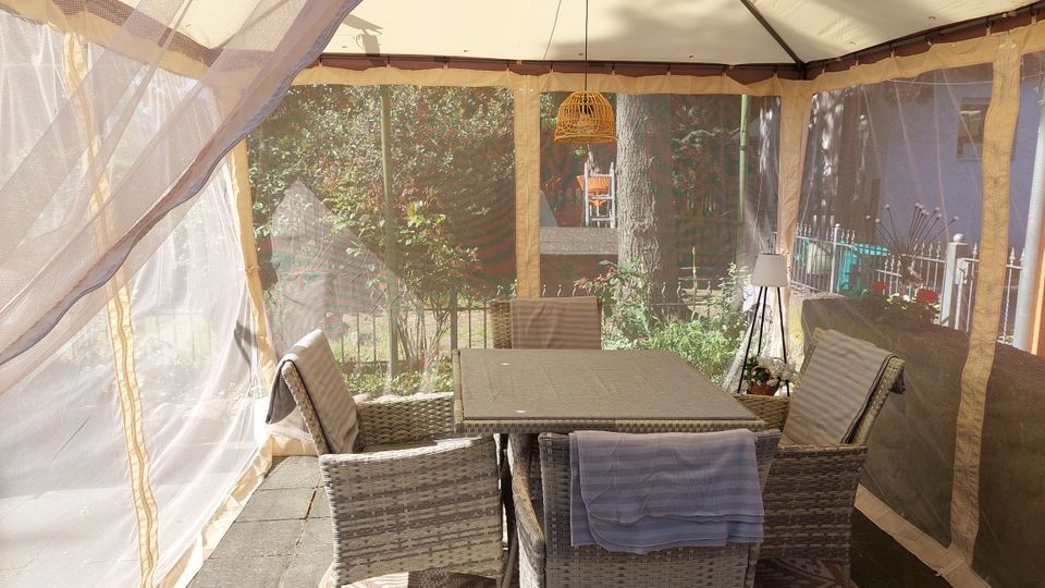 Überdachte Gartenterrasse in einem Zeltpavillon mit lichtdurchlässigen Seitenteilen, ausgestattet mit einem rechteckigen Tisch und mehreren Rattan-Sesseln. Im Hintergrund sind ein umzäunter Garten mit Bäumen und Sträuchern sowie angrenzende Wohngebäude zu sehen.