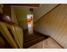 Blick in ein enges Treppenhaus mit Holzstufen, dunkelbraunem Handlauf und holzvertäfelter Wandverkleidung. Unten führt ein Flur zu einer offenen Tür in einen hellen Raum mit Fenster und Tisch.