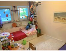 Kinderzimmer mit einem kleinen weißen Bett mit roter Bettdecke und Herzmuster-Kissen, einem Regal mit Büchern und Spielsachen sowie zwei Fenstern mit bunten Fensteraufklebern. An der Wand hängt ein Bild mit Strandmotiv, und der Boden ist teils mit Teppich bedeckt.
