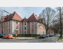 Mehrstöckiges Wohngebäude im klassischen Stil mit ziegelrotem Walmdach und einer cremefarbenen Fassade mit orangefarbenen Akzenten. Vor dem Gebäude verläuft eine gepflasterte Straße mit parkenden Autos und kahlen Bäumen.