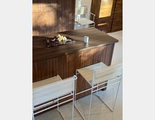 Modern kitchen island with dark wood paneling and a matching wood countertop, set on a light speckled floor. Two white wire-frame bar stools sit at the island, and a marble tray with grapes and a small cup rest on the surface, with wood cabinetry and a glass door in the background.