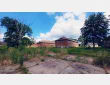 Backsteingebäude einer verlassenen Anlage mit mehreren Etagen und zahlreichen Fenstern, umgeben von überwuchertem Gras und vereinzelten Bäumen, unter einem blauen Himmel mit weißen Wolken. Im Vordergrund ist ein verfallener, betonierter Bereich zu sehen.