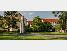 Ein kleiner Park mit einer steinernen Gedenksäule, die eine rote Sternverzierung trägt, umgeben von gepflegten Grünflächen, Bäumen und gelb gestrichenen Wohnhäusern mit roten Ziegeldächern im Hintergrund.