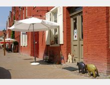 Straßenszene mit roten Backsteingebäuden, weißen Fensterläden und Türen in Beige. Vor einem Geschäft stehen dekorative Figuren, und ein weißer Sonnenschirm spendet Schatten; im Hintergrund führen weitere Gebäude und Sitzmöglichkeiten entlang des gepflasterten Gehwegs.
