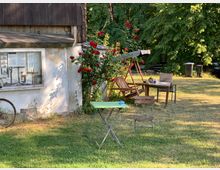 Ein Garten mit einer kleinen, alten Hütte mit weißen Wänden und dunklem Holzdach. Vor der Hütte steht ein bepflanzter Rosenstrauch, daneben Gartenmöbel, darunter ein Tisch, Stühle und eine Holzschaukelbank, auf einer Wiese umgeben von Bäumen.