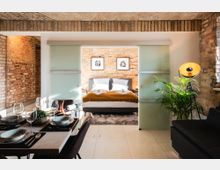 Loft-Wohnung mit Backsteinwänden und modernem Design: Im Vordergrund ein Esstisch mit gedeckten Plätzen, dahinter ein Schlafzimmer mit Doppelbett, das durch mattierte Glasschiebetüren abgetrennt ist. Eine Stehlampe und eine Zimmerpflanze ergänzen die Einrichtung.