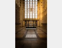 Große, hohe Eingangshalle mit einem breiten Bodenmuster aus dunklen und hellen Steinfliesen, großen Säulen an den Seiten und einer Fensterfront mit Buntglas im Hintergrund. Im Zentrum steht ein rechteckiger, niedriger Infotisch.