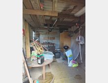 Innenansicht einer einfachen Garage bzw. eines Schuppens mit sichtbaren Holzbalken an der Decke, Betonwänden und einem gemusterten Bodenbelag. Der Raum ist mit gelagerten Gegenständen wie Tisch, Werkzeugbank, Schrank, Säcken, Kanistern, Brettern und einem Kugelgrill vollgestellt.