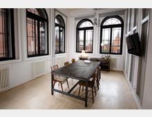 Heller Besprechungsraum mit einem großen Holztisch und verschiedenen Stühlen in der Mitte, umgeben von weißen Wänden mit großen, halbkreisförmigen schwarzen Fensterrahmen, die Ausblick auf Backsteinbauten bieten. Heizkörper befinden sich unter den Fenstern, und ein Regal mit Getränken steht im Hintergrund.