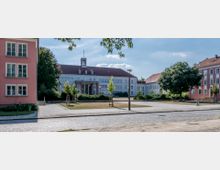 Hauptplatz mit einem großen, symmetrischen Gebäude in der Mitte, das eine weiße Fassade, ein rotes Dach und einen kleinen Uhrturm besitzt. Umgeben von weitläufigen Grünflächen, gepflasterten Gehwegen sowie rosa und weißen Wohnhäusern im Hintergrund.