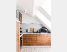 Moderne Küche mit Holzfronten unter einer Dachschräge. Sichtbar sind ein Herd, eine Spüle, eine Arbeitsplatte mit Küchenutensilien und ein Mikrowellenofen; der Boden besteht aus hellem Holz.