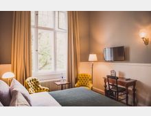 Helles Schlafzimmer mit grauen Wänden, einem großen Fenster mit beigefarbenen Vorhängen und einem Bett im Vordergrund. Der Raum ist ausgestattet mit zwei gelben Polstersesseln, einem kleinen Holztisch, einer Stehlampe, einem Schreibtisch, einem Stuhl und einem flachen Fernseher an der Wand.