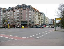 Straßenszene in einem urbanen Gebiet mit Wohn- und Geschäftsgebäuden im Hintergrund, darunter ein markantes mehrstöckiges Gebäude mit abgerundeten Fenstern. Im Vordergrund befinden sich eine breite Kreuzung mit markierten Fußgängerüberwegen, parkende Autos und ein Wegweiser mit dem Schriftzug „Wedding“.
