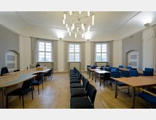 Großer Besprechungsraum mit Parkettboden, einer halbkreisförmigen Theke aus Holz auf der linken Seite, Stuhlreihen in der Mitte und rechteckigen Tischen mit blauen Stühlen rechts. Der Raum hat hohe Fenster, helle Wände mit vertikalen Verzierungen und eine moderne Hängelampe aus Metall und Glas.