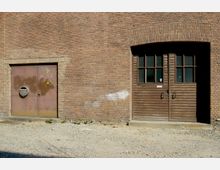 Ziegelsteinfassade eines Gebäudes mit zwei Türen: links eine rostige Metalltür mit rundem Lüftungsgitter, rechts eine doppelflüglige Holztür mit Glasfenstern und bogenförmigem Mauerwerk darüber. Der Boden davor besteht aus Schotter.