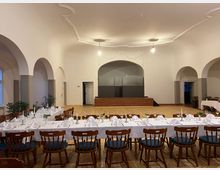 Ein festlich gedeckter Saal mit langen, weiß eingedeckten Tischen und Holzstühlen mit blauen Sitzpolstern. Der Raum hat helle Wände, hohe Decken mit einfacher Stuckverzierung und mehrere Rundbogenöffnungen; im Hintergrund befindet sich eine kleine Holzbühne.