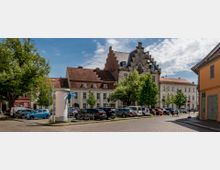 Städtischer Platz mit Kopfsteinpflaster, umgeben von historischen Gebäuden mit roten Ziegeldächern und Giebelarchitektur. Im Vordergrund sind geparkte Autos, Fahrräder und vereinzelte Bäume zu sehen.