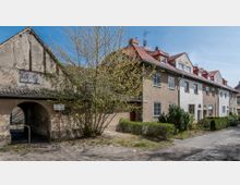 Reihenhäuser mit roten Ziegeldächern und hellen Fassaden in einer Wohnsiedlung, daneben ein älteres Gebäude mit verputzter Fassade und einem Rundbogendurchgang. Davor stehen ein Baum mit frischem Laub und Büsche, im Hintergrund blauer Himmel.