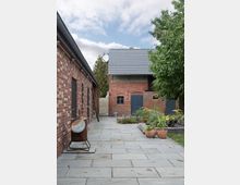 Gepflasterter Innenhof zwischen Backsteingebäuden, mit Pflanzkübeln, einem Baum und einem Fass auf einem Ständer; im Hintergrund ein Gebäude mit grauem Ziegeldach und blauen Türen.