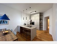 Moderne Küche mit weißer Einbauküche, integrierten Geräten und einer zentralen Kücheninsel mit grauer Arbeitsplatte und zwei Barhockern. Der Raum hat einen Holzboden, weiße Wände und minimalistische Hängelampen; ein Esstisch mit Holzplatte steht im Vordergrund.