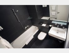 Modernes Badezimmer mit dunklen Fliesenwänden, einer weißen Badewanne mit Handbrause, einem weißen Waschbecken mit Spiegel und einem wandhängenden WC. Im Hintergrund ein Handtuchheizkörper und ein kleines Regal mit Glasablage.