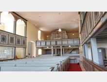 Innenansicht einer historischen Kirche mit hell gestrichenen Holzbänken, einer Empore mit bemalten Tafeln und Schriftzügen sowie einer Orgel im Hintergrund. Die Wände sind in hellem Beige gehalten, und es sind dekorative Deckenlampen zu sehen.