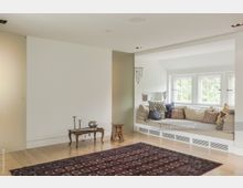 Wohnbereich mit minimalistischem Design, hellem Holzboden, einer großen Sitzbank mit Kissen unter bodentiefen Fenstern und einem orientalischen Teppich im Vordergrund. Ein kleiner Holztisch mit Kerzen und Dekoration steht an einer weißen Wand.