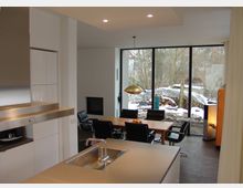 Offener Wohnbereich mit moderner Küche im Vordergrund, bestehend aus einer Arbeitsfläche mit Spüle. Im Hintergrund ein Esszimmer mit Holztisch und schwarzen Stühlen vor einer großen Fensterfront mit Blick auf einen verschneiten Garten.