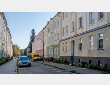 Eine schmale Wohnstraße mit mehrstöckigen, pastellfarbenen Altbauten auf beiden Seiten; geparkte Autos und ein gelber Lieferwagen befinden sich am Straßenrand. Im Hintergrund sind Bäume und ein klarer blauer Himmel zu sehen.