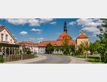 Straßenszene in einer kleinen Stadt mit historischen Gebäuden, darunter eine Kirche mit rotem Ziegeldach und einem schwarzen Turm in der Mitte. Rechts ein auffälliges gelbes Gebäude mit barocken Elementen, umgeben von Bäumen und Grünflächen.